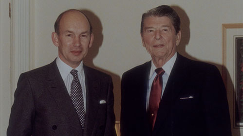 Two men in suits stand side by side indoors, both facing the camera and smiling slightly. One wears a dark polka-dot tie, and the other wears a red tie. They are standing in front of a light-colored wall with framed artwork.