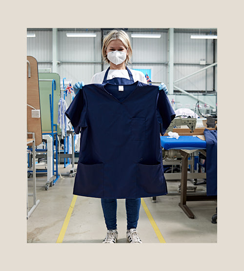 A person wearing a mask, gloves, and casual clothes stands in a factory setting, holding up a large navy blue scrub top. Sewing machines and workstations are visible in the background.