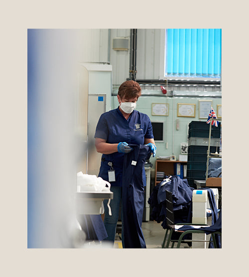 A person wearing blue scrubs, gloves, and a face mask stands in a workshop handling fabric with sewing machines and a small British flag in the background. Blue blinds and certificates are visible on the wall.