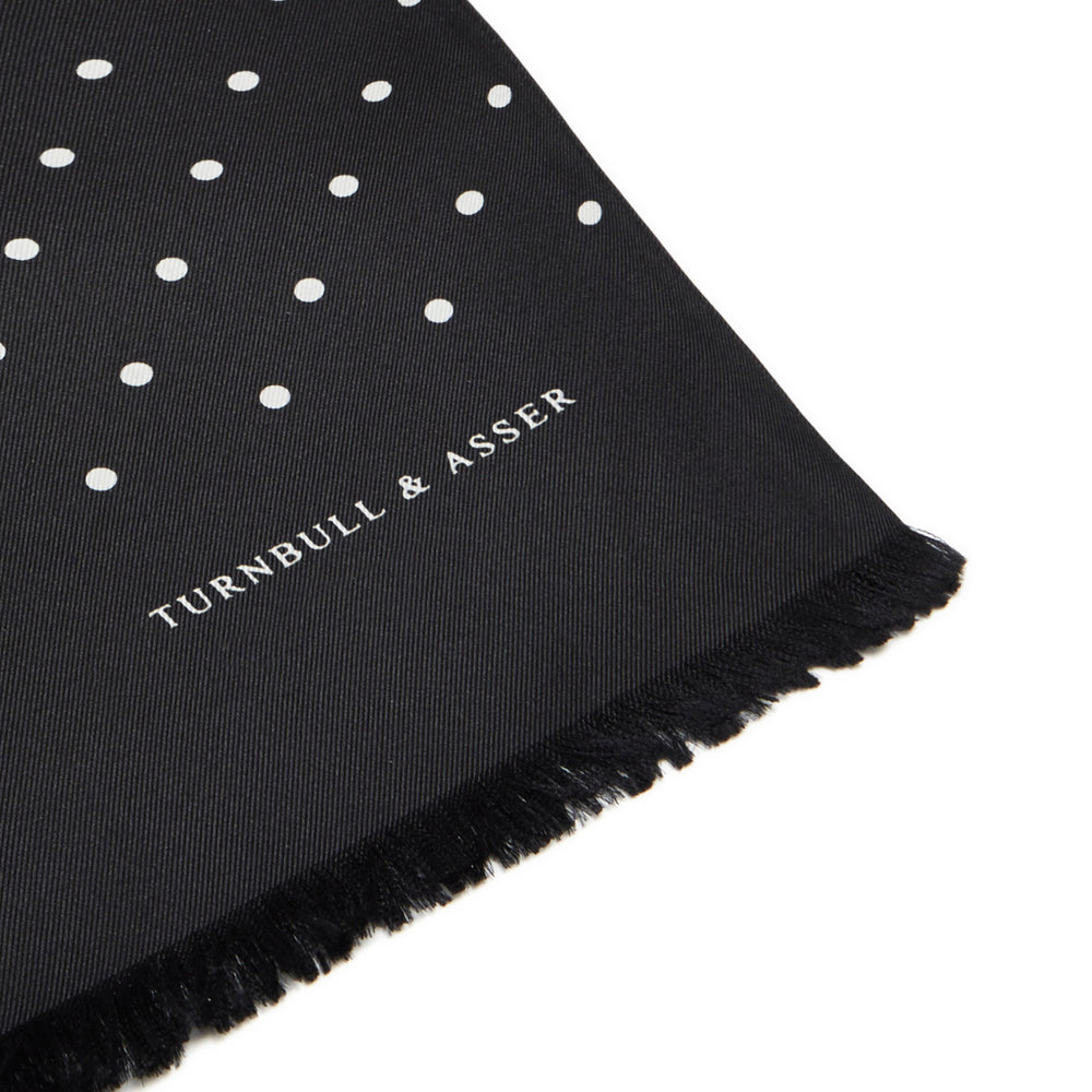 The Black and White Spotted Silk Scarf by Turnbull & Asser features small hand-printed white polka dots and short fringes, shown laid flat on a white background.