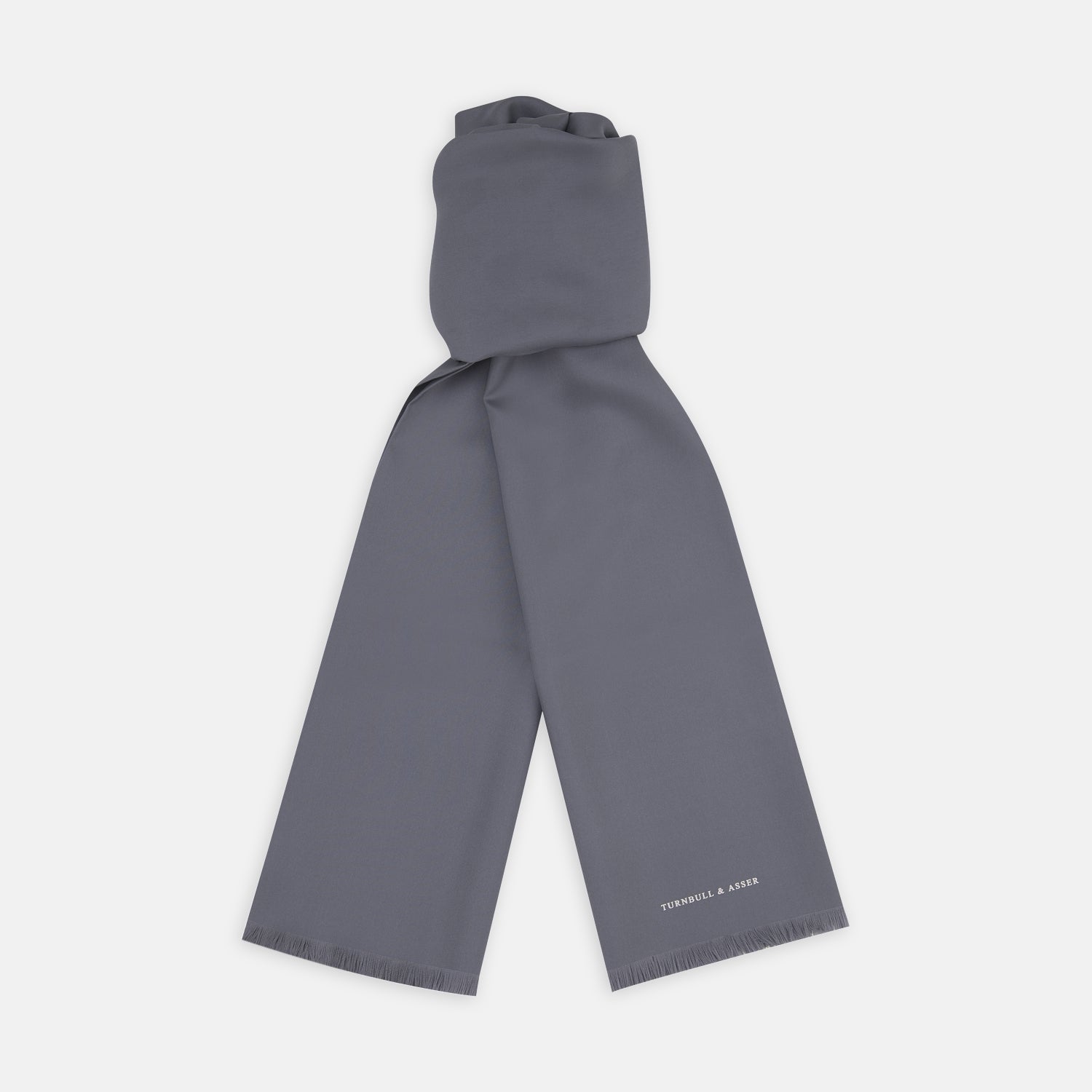 View 1 of product The Plain Grey Silk Scarf by Turnbull & Asser features fringed edges and the brand name in white near one end, neatly folded and elegantly displayed on a plain white background.