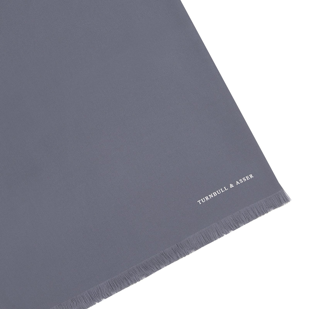 The Plain Grey Silk Scarf by Turnbull & Asser features fringed edges and the brand name in white near one end, neatly folded and elegantly displayed on a plain white background.