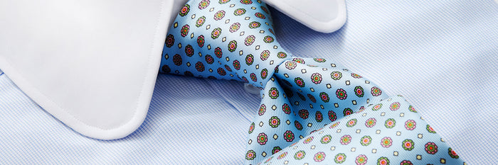 Close-up of a light blue dress shirt with a white collar, paired with a light blue patterned necktie featuring small red and yellow floral designs and white diamond shapes.