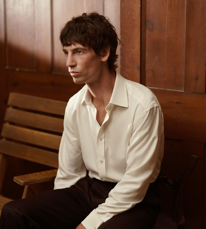A man with medium-length dark hair, wearing a white dress shirt and dark pants, sits on a wooden bench in front of wooden paneling, looking slightly to the side with a neutral expression.