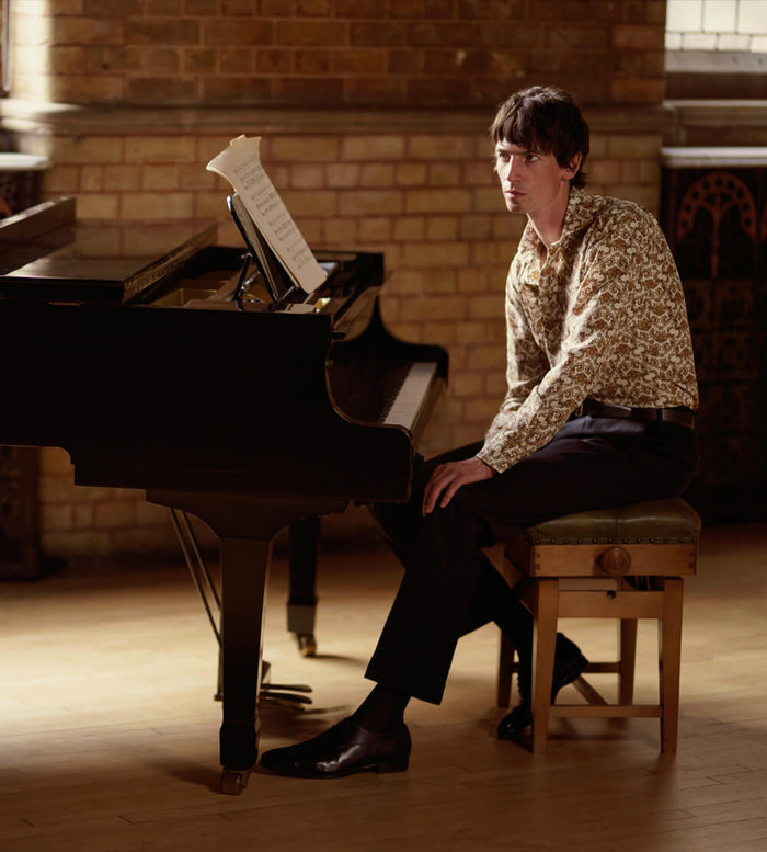 A man in a patterned shirt and dark pants sits on a bench at a grand piano, looking away from the keys, in a sunlit room with brick walls. Sheet music rests on the piano.