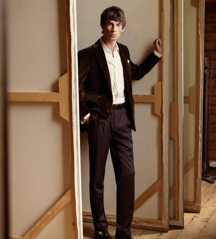 A man in a dark suit and cream shirt stands indoors, leaning against a wooden frame with one hand in his pocket, looking at the camera. The setting appears artistic, with large framed canvases behind him.