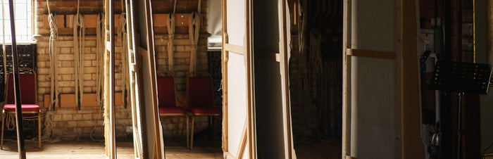 Several tall, narrow wooden panels stand upright in a dimly lit room with brick walls, ropes, red chairs, and a window letting in natural light.