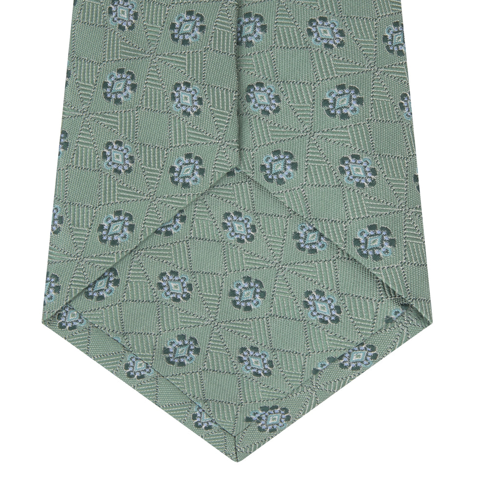 The Turnbull & Asser Green Star Flowers Silk Tie features a light green jacquard with a subtle checkered pattern and small blue star flower motifs, shown rolled up on a white background.