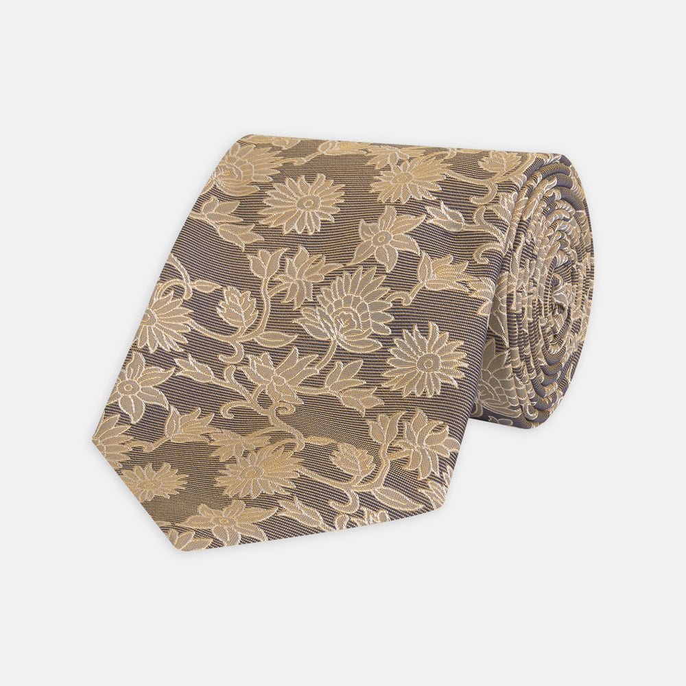 The Copper Floral Occasion Silk Tie by Turnbull & Asser features a gold floral jacquard pattern on brown silk, elegantly rolled on a white surface—an ideal choice for weddings.