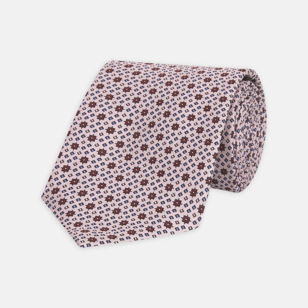 The Turnbull & Asser Pastel Pink Geometric Silk Tie features a small, repeating dark brown and white pattern for a mid-century look—ideal with tailored jackets. Shown rolled up on a white background.