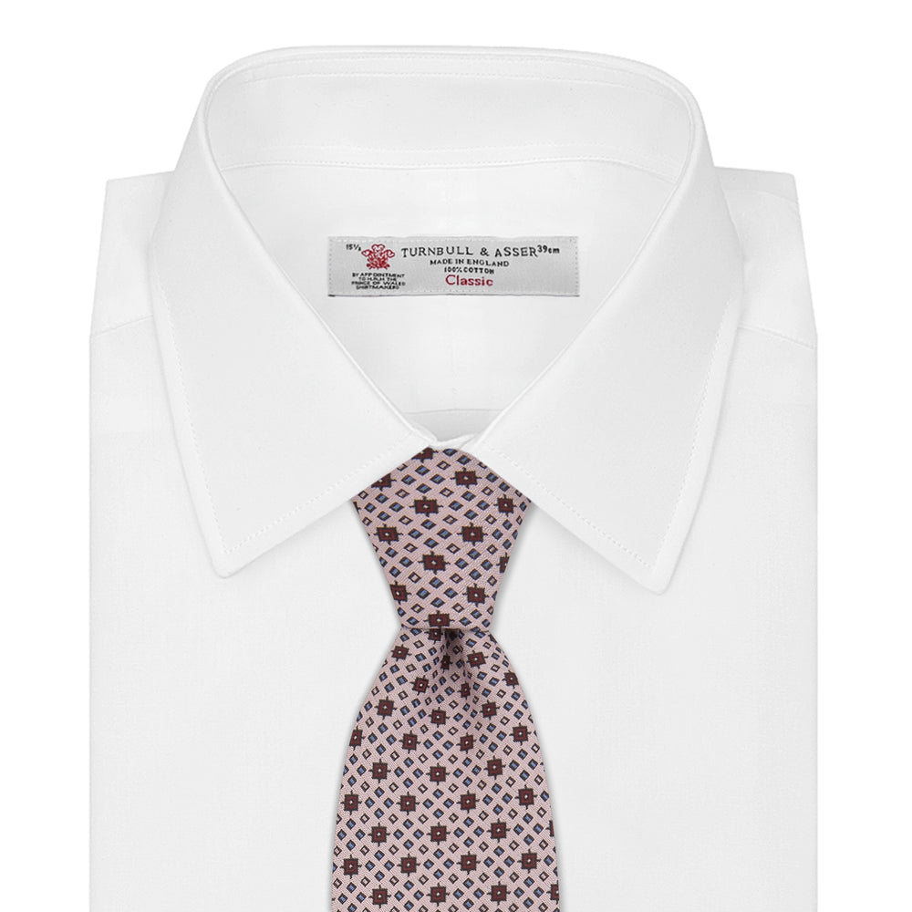 The Turnbull & Asser Pastel Pink Geometric Silk Tie features a small, repeating dark brown and white pattern for a mid-century look—ideal with tailored jackets. Shown rolled up on a white background.