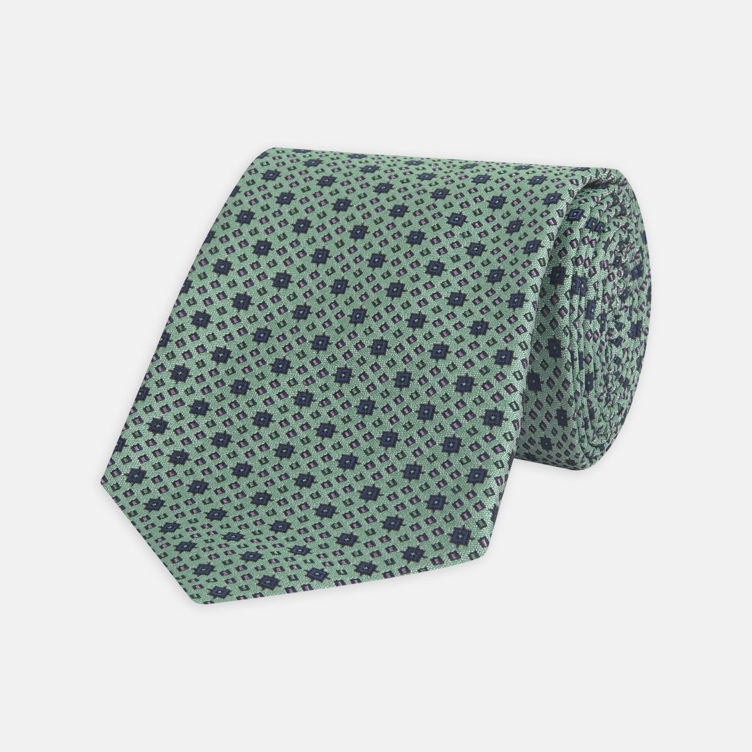 View 1 of product The Leaf Green Geometric Silk Tie by Turnbull & Asser features a repeating pattern of small navy blue and white floral shapes, evoking a mid-century motif. It is neatly rolled and displayed on a white background.