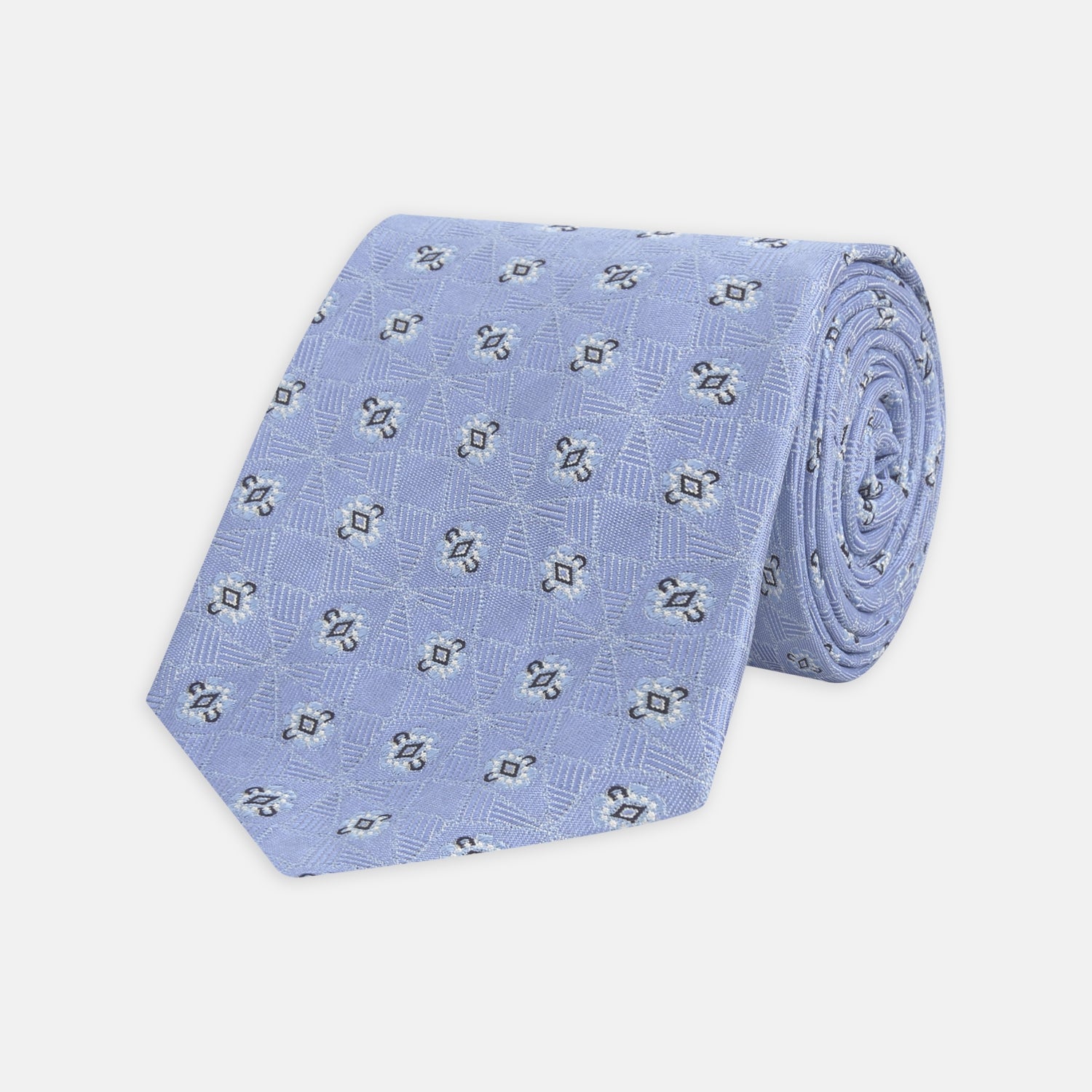 View 1 of product The Turnbull & Asser Sky Blue Star Flowers Silk Tie features a small white and dark blue square and star flower pattern on jacquard silk, shown rolled up against a plain white background.