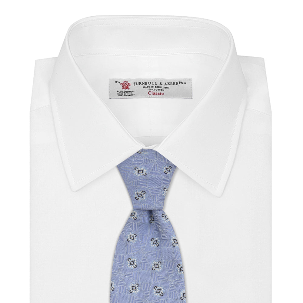 The Turnbull & Asser Sky Blue Star Flowers Silk Tie features a small white and dark blue square and star flower pattern on jacquard silk, shown rolled up against a plain white background.