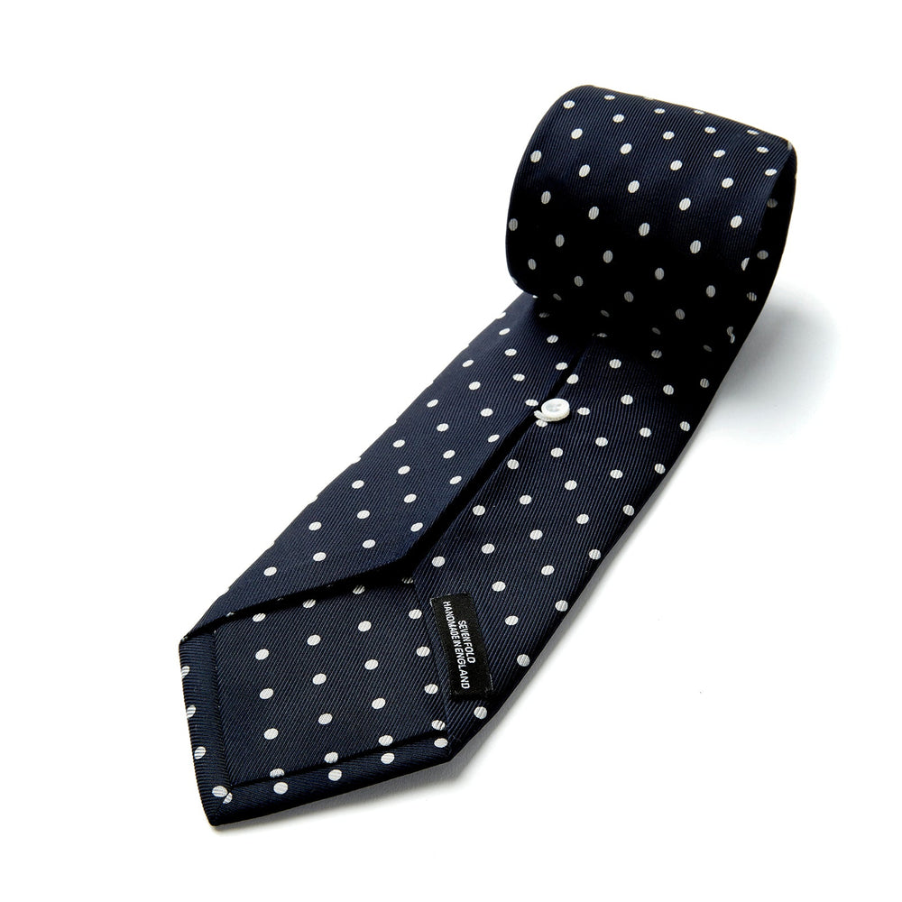 The Seven-Fold Navy Churchill Spot Silk Tie by Turnbull & Asser features evenly spaced white polka dots on navy silk—a luxury neckwear piece rolled up and displayed on a white background.