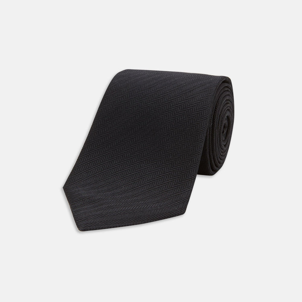 The Turnbull & Asser Black Herringbone Silk Tie is rolled up and displayed on a plain white background, showcasing its classic style and subtle chevron pattern.