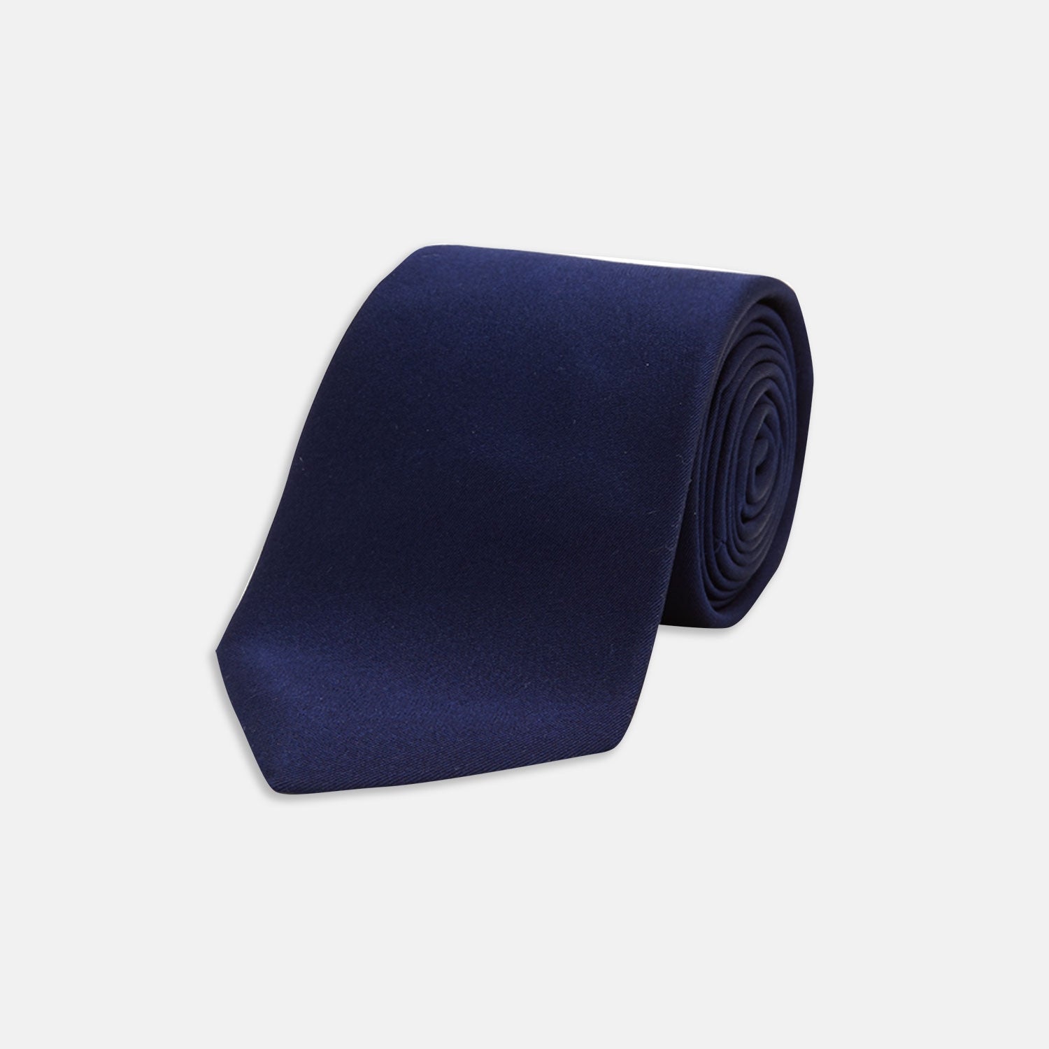 View 1 of product The Turnbull & Asser Seven-Fold Navy Plain Satin Silk Tie is neatly rolled and showcased against a plain light gray background.