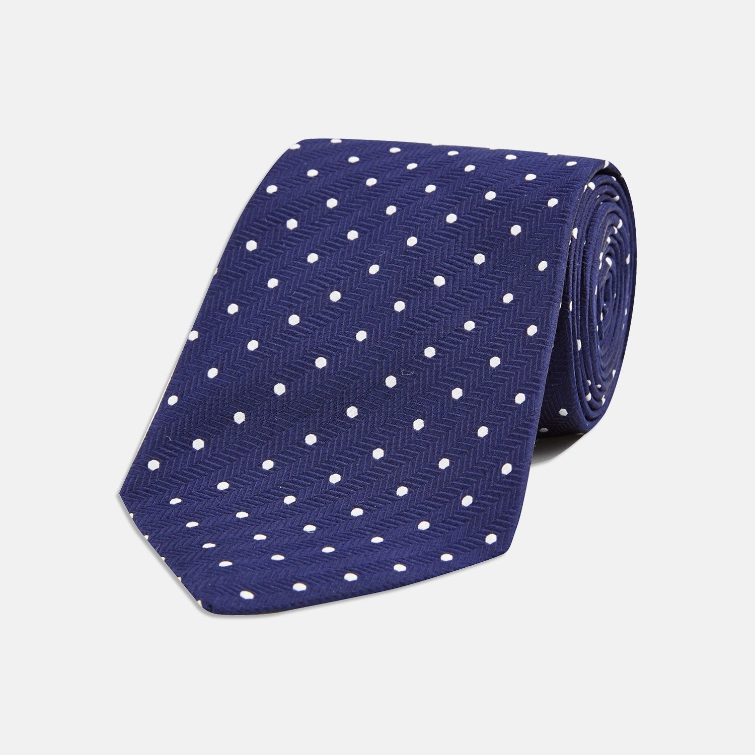 View 1 of product The Turnbull & Asser Seven-Fold Navy & White Spot Herringbone Silk Tie features a navy base with white polka dots and a classic herringbone weave, displayed rolled up on a plain white background.