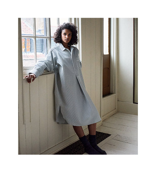 A woman with curly hair wearing a loose, light blue striped shirt dress leans against a white-paneled wall in a bright room with large windows and wooden floors.