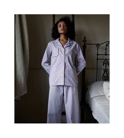 A person stands confidently in a dimly lit bedroom wearing blue-and-white striped pajamas, next to an ornate metal bed with white bedding.
