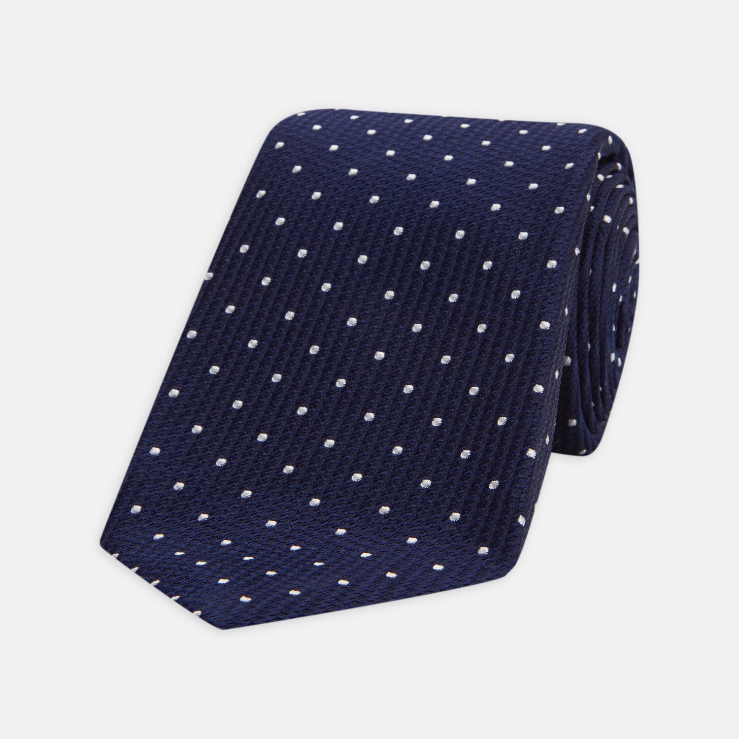 View 1 of product The Seven-Fold Navy and White Spot Lace Silk Tie by Turnbull & Asser showcases a luxurious navy silk with white spots, elegantly rolled and presented on a white background.