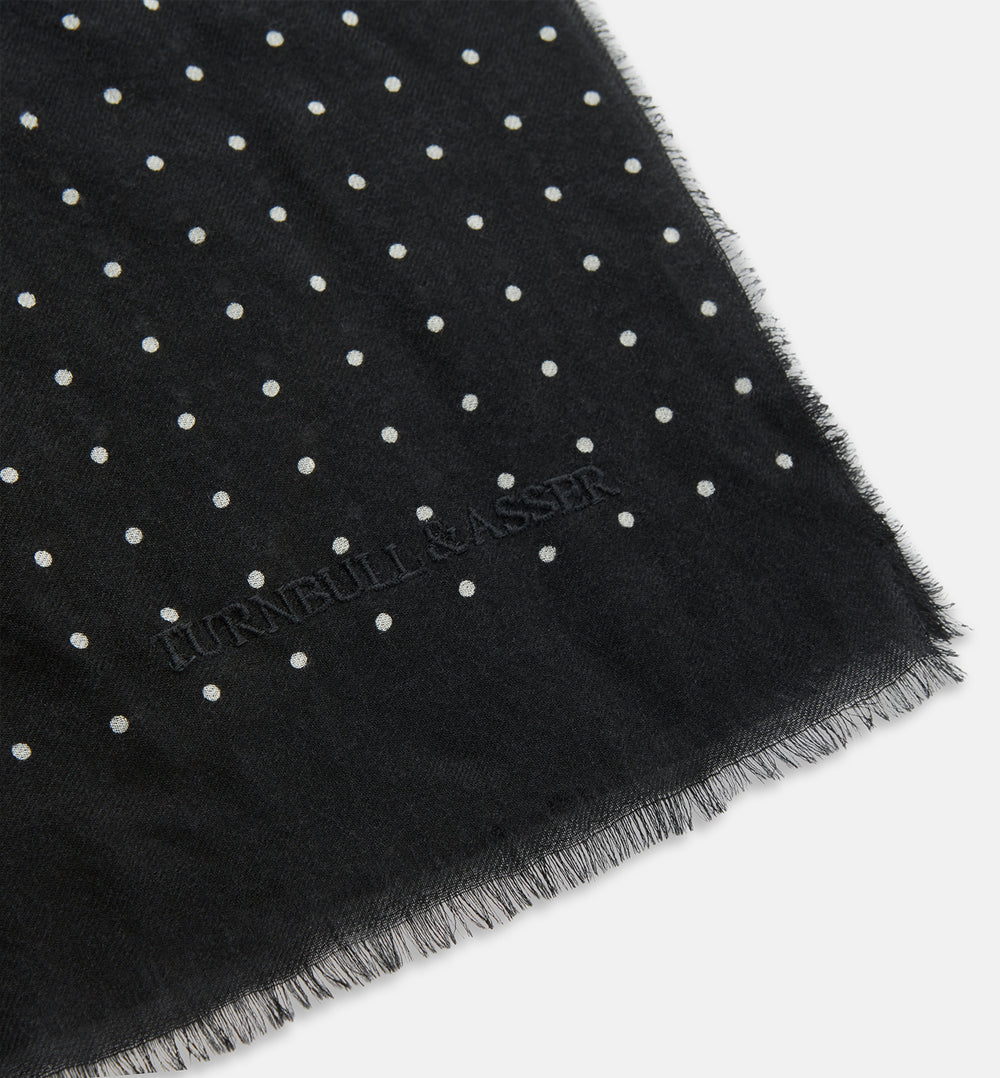 The Turnbull & Asser Black & White Spot Superfine Cashmere Scarf features evenly spaced white dots, lightly frayed edges, and a lightweight design for subtle elegance in any outfit. Shown laid flat with a loose knot at the top.