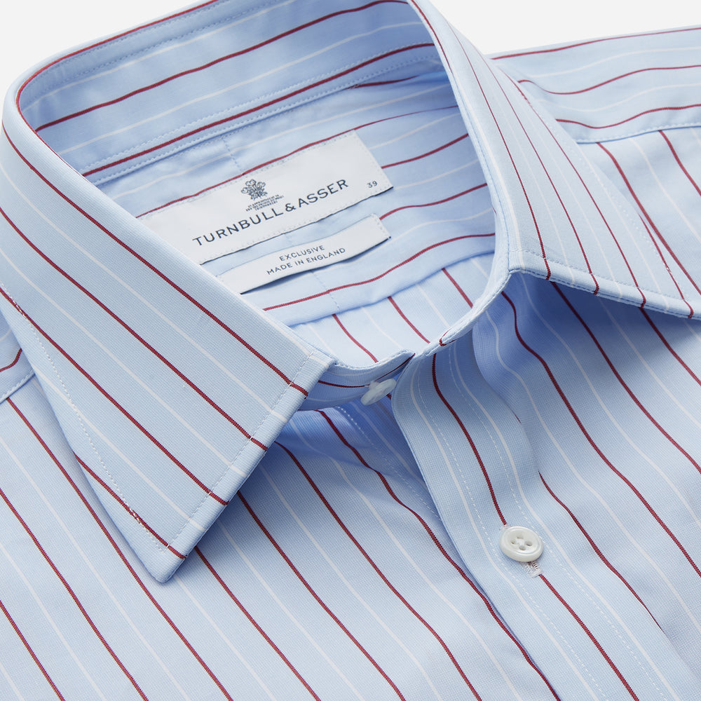 The Turnbull & Asser Burgundy Stripe Regular Fit Shirt features a T&A collar, double cuffs, and white buttons. Made from light blue Italian cotton poplin with vertical burgundy and white pinstripes. Laid flat on a white background.
