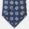 View 4 of product The Turnbull & Asser Blue Geometric Jacquard Tie is a rolled-up silk tie showcasing a repeating light blue and white geometric floral pattern with dotted accents, set against a plain light background.