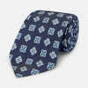 View 1 of product The Turnbull & Asser Blue Geometric Jacquard Tie is a rolled-up silk tie showcasing a repeating light blue and white geometric floral pattern with dotted accents, set against a plain light background.