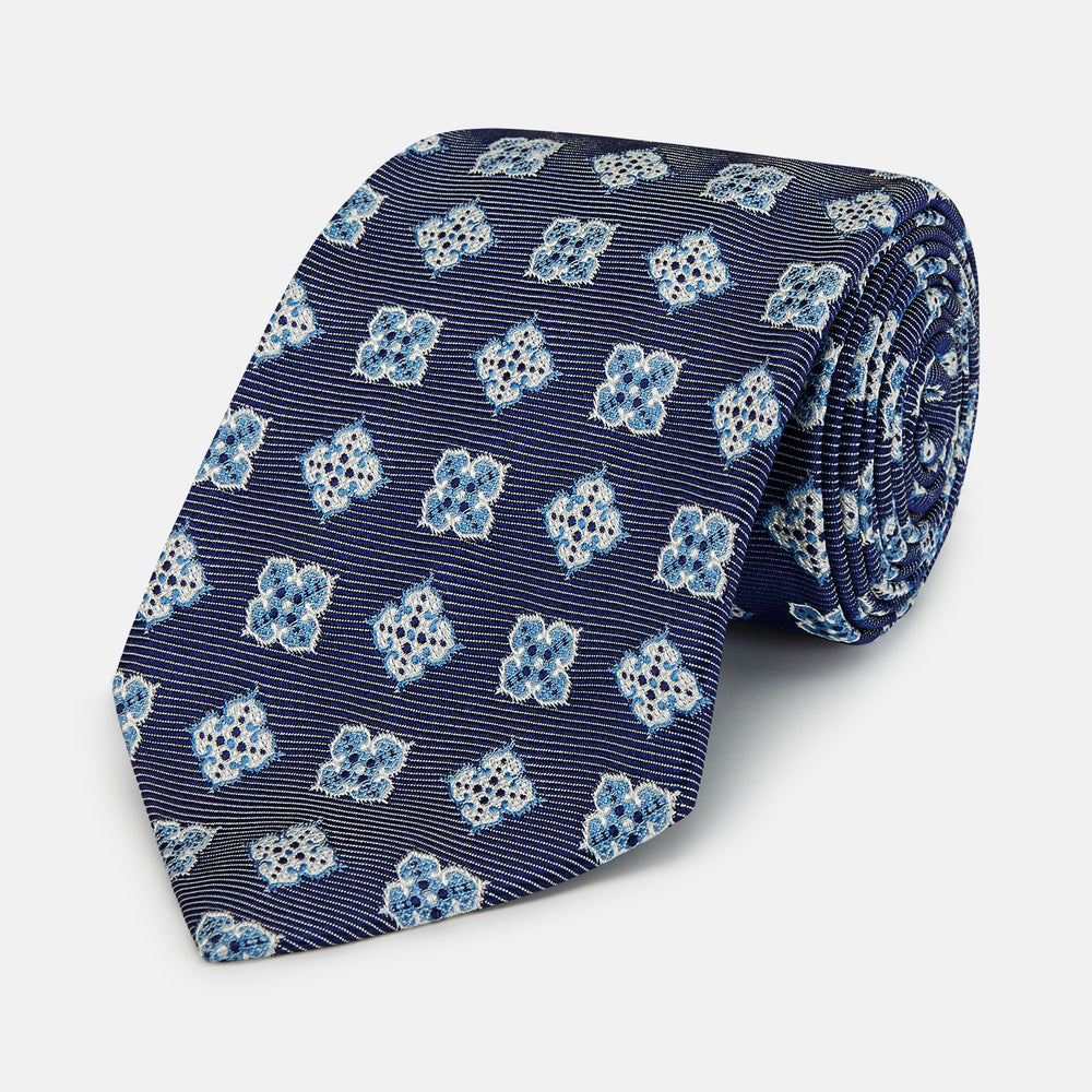 The Turnbull & Asser Blue Geometric Jacquard Tie is a rolled-up silk tie showcasing a repeating light blue and white geometric floral pattern with dotted accents, set against a plain light background.