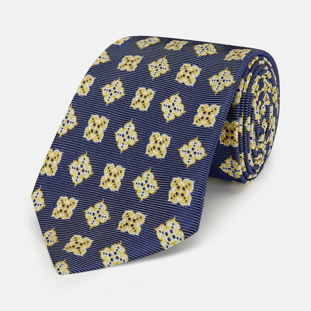 The Turnbull & Asser Yellow Geometric Jacquard Tie in navy features a repeating pattern of yellow geometric shapes with blue accents, presented rolled up on a white background.