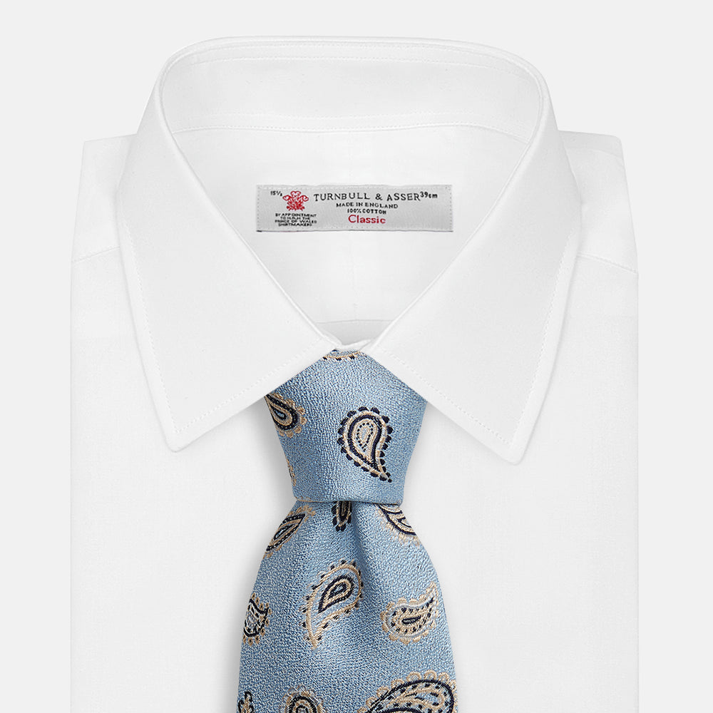 The Turnbull & Asser Sky Blue Renoir Paisley Tie, crafted in silk with a repeating navy and beige pattern, is shown rolled up with its pointed end forward on a plain white background.