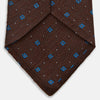 View 4 of product The Turnbull & Asser Brown Square Silk Tie, featuring a jacquard pattern of small blue squares and dots, is shown rolled up on a plain white background.