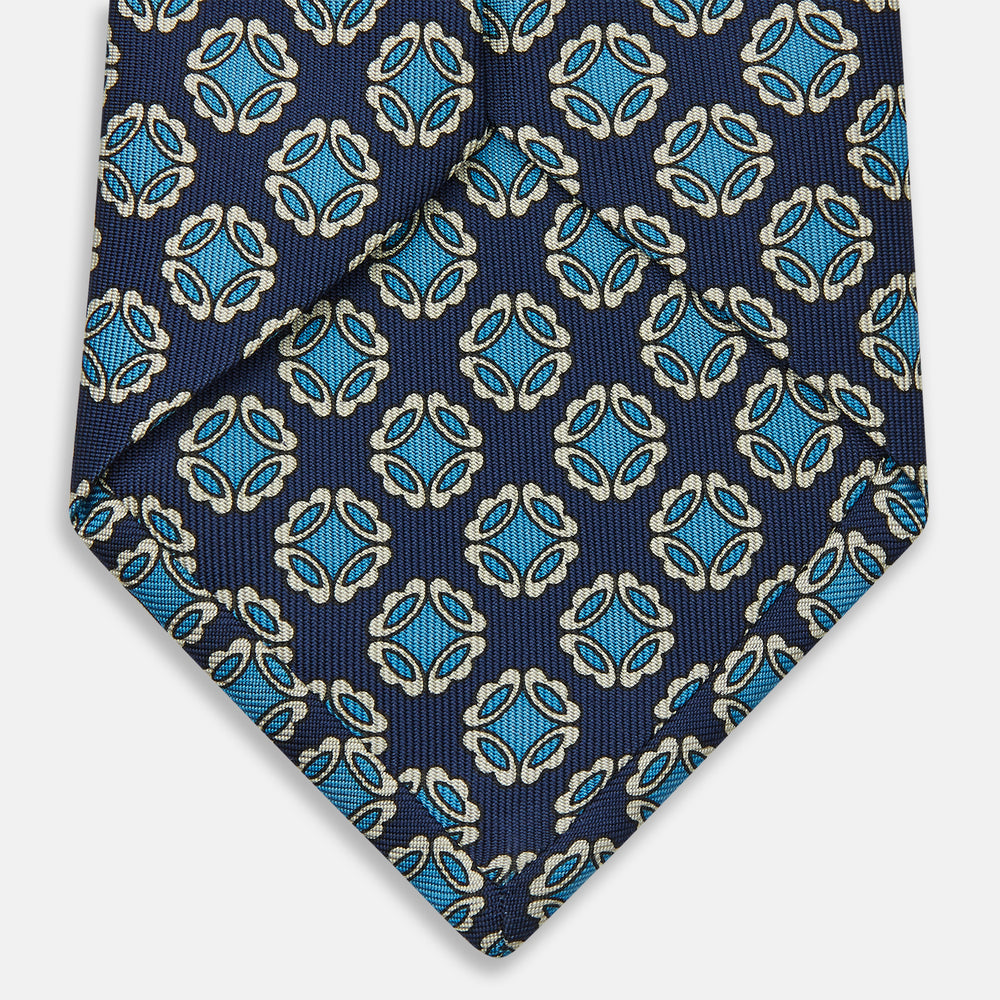 A rolled Navy Floral Geometric Tie by Turnbull & Asser, shown on a light background with light blue and white geometric floral motifs—a refined, stylish accessory.