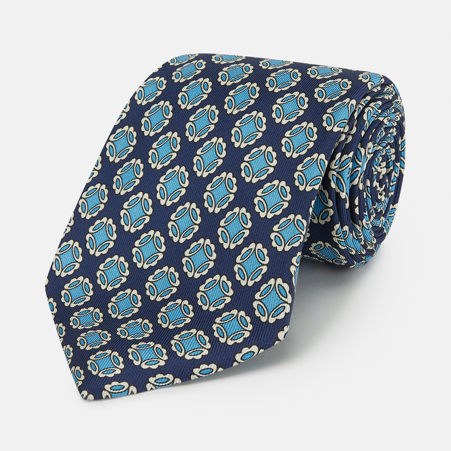 View 1 of product A rolled Navy Floral Geometric Tie by Turnbull & Asser, shown on a light background with light blue and white geometric floral motifs—a refined, stylish accessory.