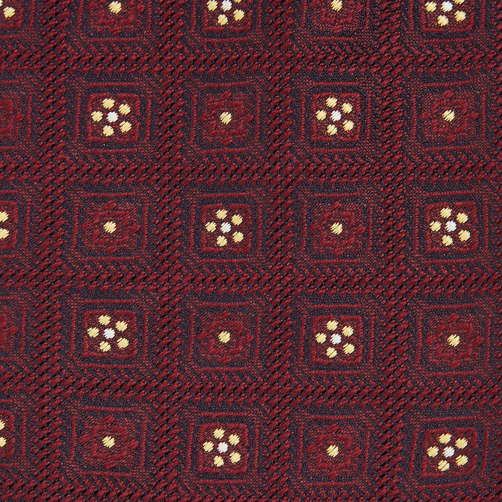 The Turnbull & Asser Burgundy Illusion Flower Silk Tie features a geometric pattern of small yellow and white dots within diamond shapes, elegantly displayed rolled up on a light background.