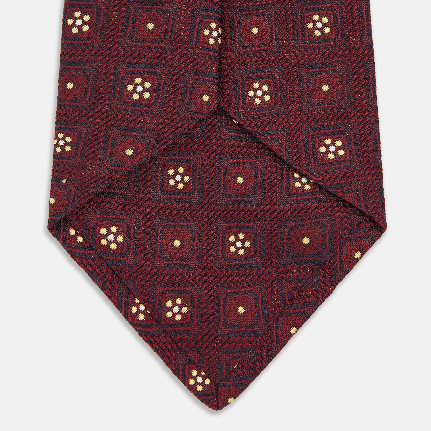 View 4 of product The Turnbull & Asser Burgundy Illusion Flower Silk Tie features a geometric pattern of small yellow and white dots within diamond shapes, elegantly displayed rolled up on a light background.