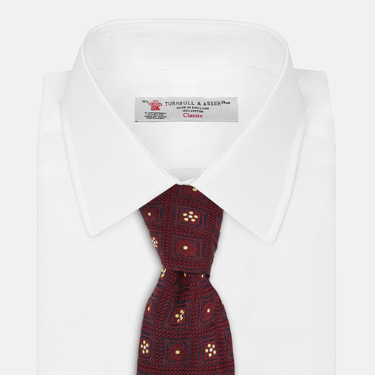 View 2 of product The Turnbull & Asser Burgundy Illusion Flower Silk Tie features a geometric pattern of small yellow and white dots within diamond shapes, elegantly displayed rolled up on a light background.