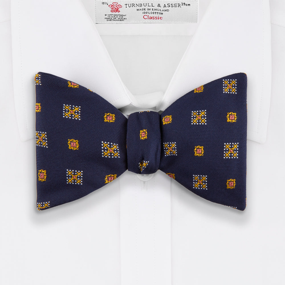 The Yellow Shields Bow Tie by Turnbull & Asser features a navy blue base with small yellow, orange, and white geometric squares, ideal for formal occasions and shown against a white background.