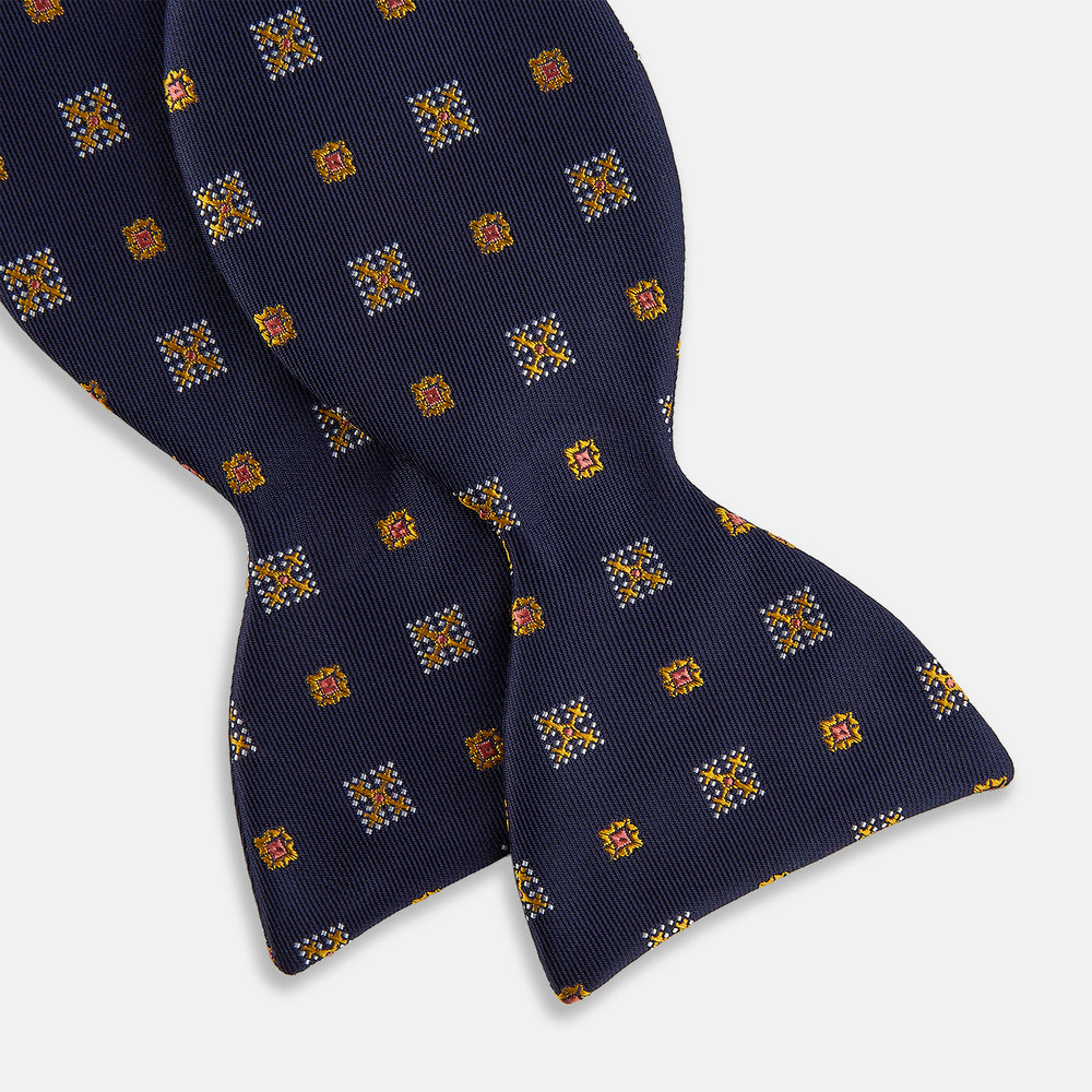 The Yellow Shields Bow Tie by Turnbull & Asser features a navy blue base with small yellow, orange, and white geometric squares, ideal for formal occasions and shown against a white background.