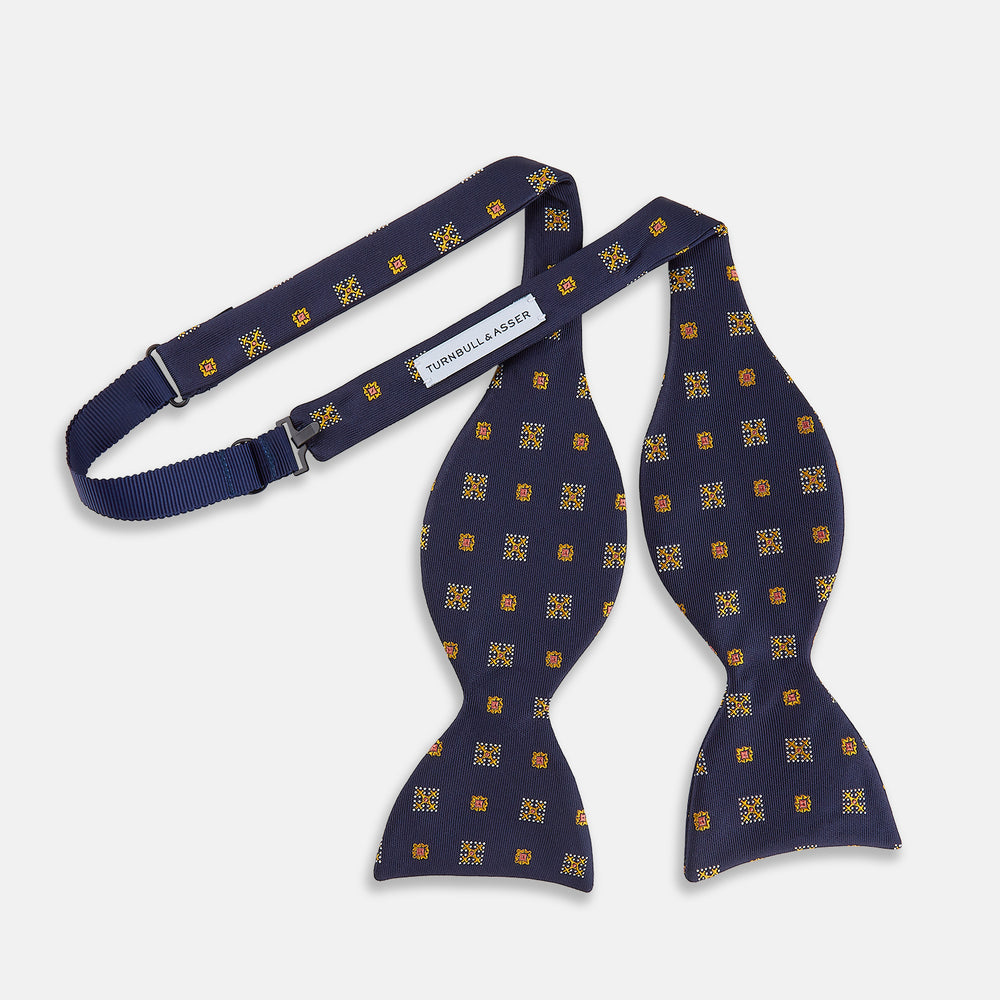The Yellow Shields Bow Tie by Turnbull & Asser features a navy blue base with small yellow, orange, and white geometric squares, ideal for formal occasions and shown against a white background.