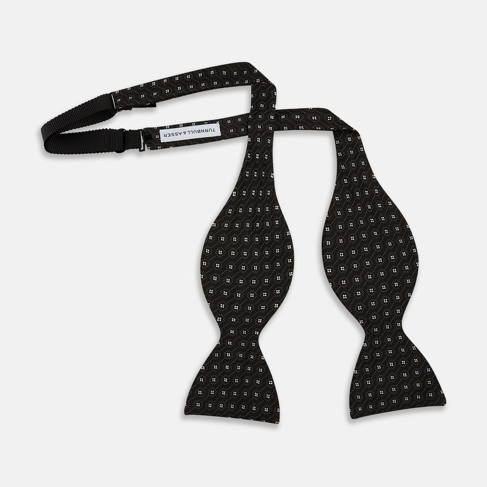 The Floral Piqué Monochrome Silk Bow Tie by Turnbull & Asser, featuring a repeating pattern of small white dots and hexagons on black silk, is ideal for formalwear or a dinner suit and is shown against a plain white background.