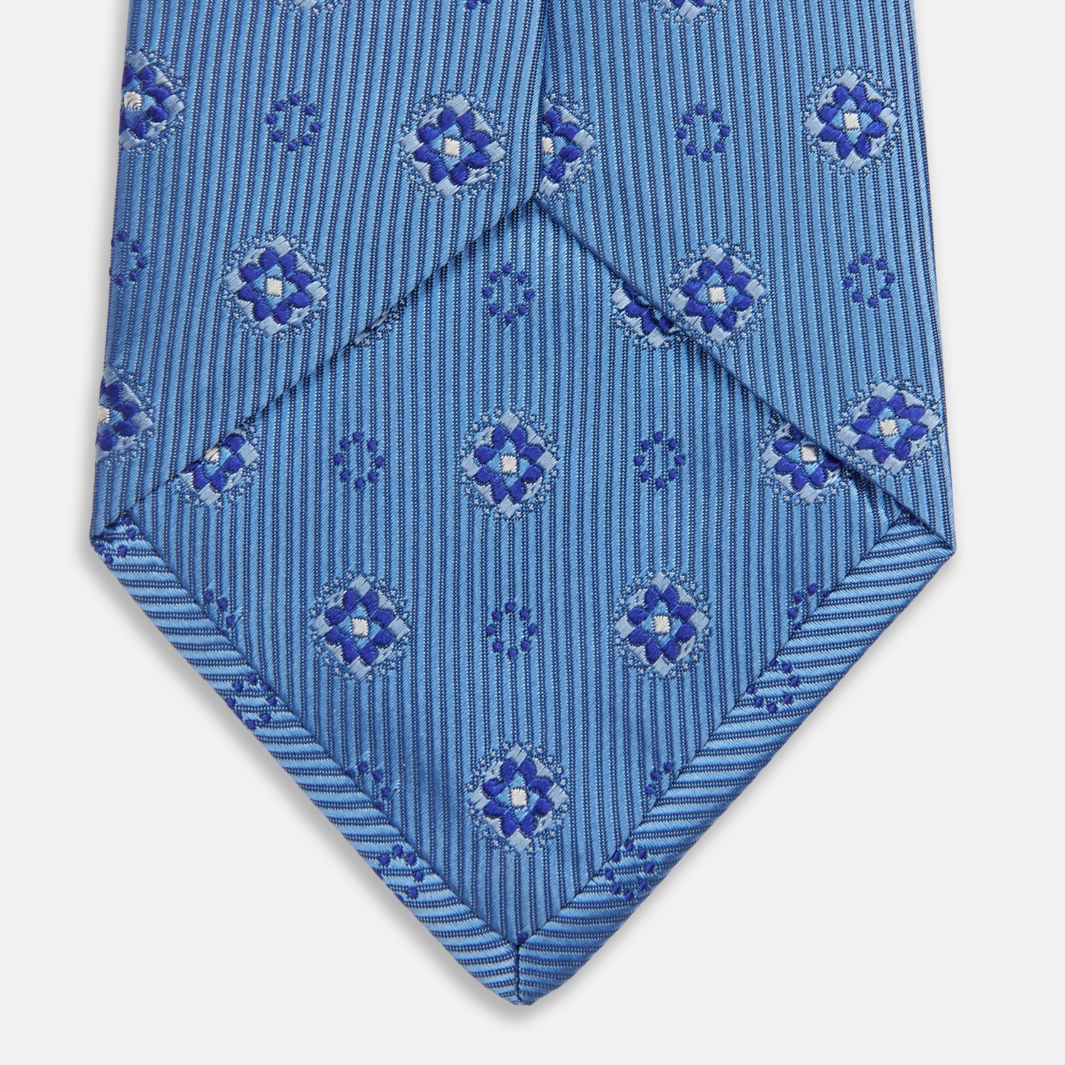 View 4 of product The Blue Multi Floral Motif Silk Tie by Turnbull & Asser features subtle diagonal stripes and blue and white floral medallions on light blue silk—ideal for bold shirts. The tie is shown rolled at one end.
