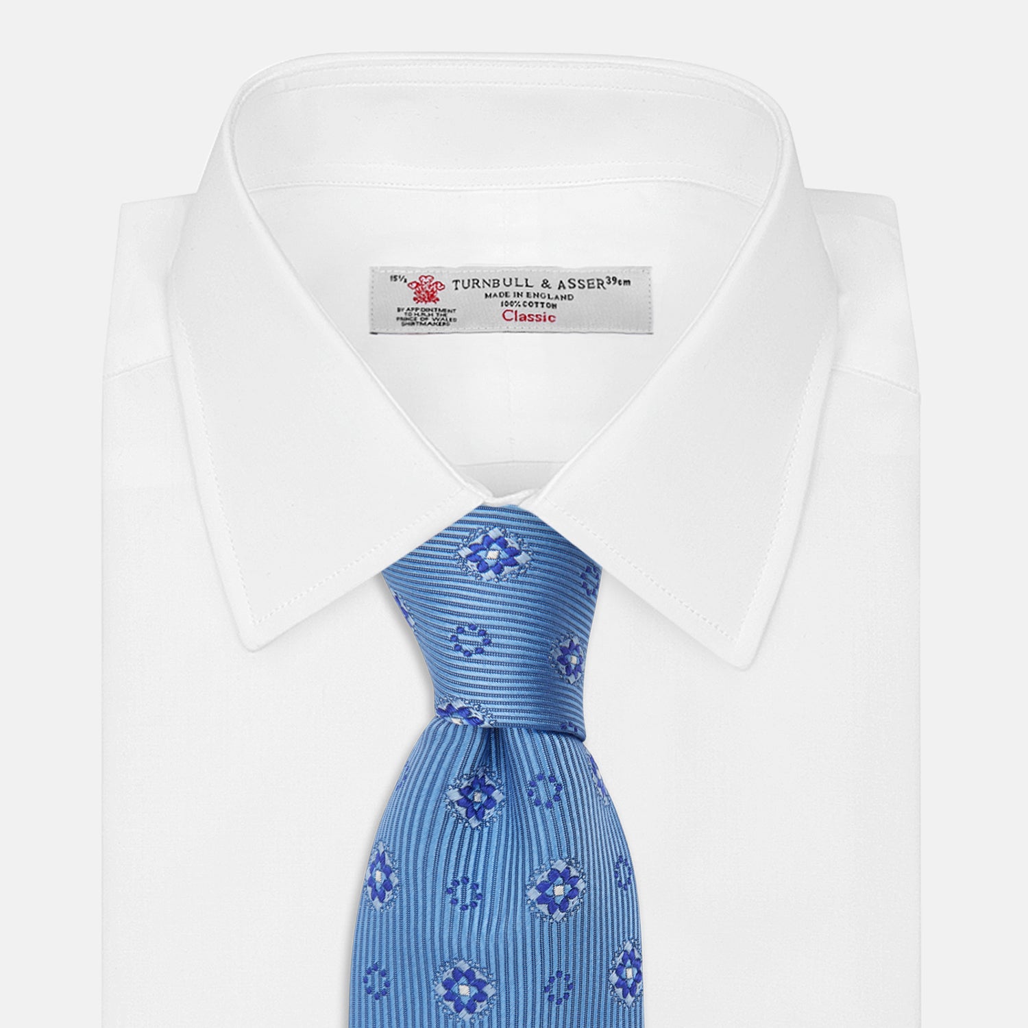 View 2 of product The Blue Multi Floral Motif Silk Tie by Turnbull & Asser features subtle diagonal stripes and blue and white floral medallions on light blue silk—ideal for bold shirts. The tie is shown rolled at one end.