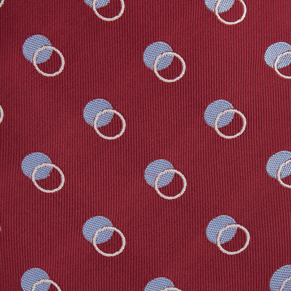 The Turnbull & Asser Red Multi Circle Silk Tie features small blue and white rings on red silk, making it ideal for pairing with bold shirts or adding vibrant flair to summer looks. Shown rolled at one end against a plain white background.