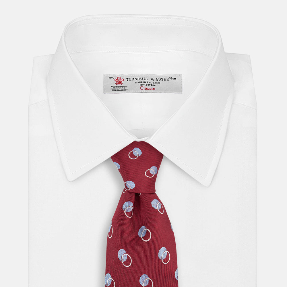 The Turnbull & Asser Red Multi Circle Silk Tie features small blue and white rings on red silk, making it ideal for pairing with bold shirts or adding vibrant flair to summer looks. Shown rolled at one end against a plain white background.