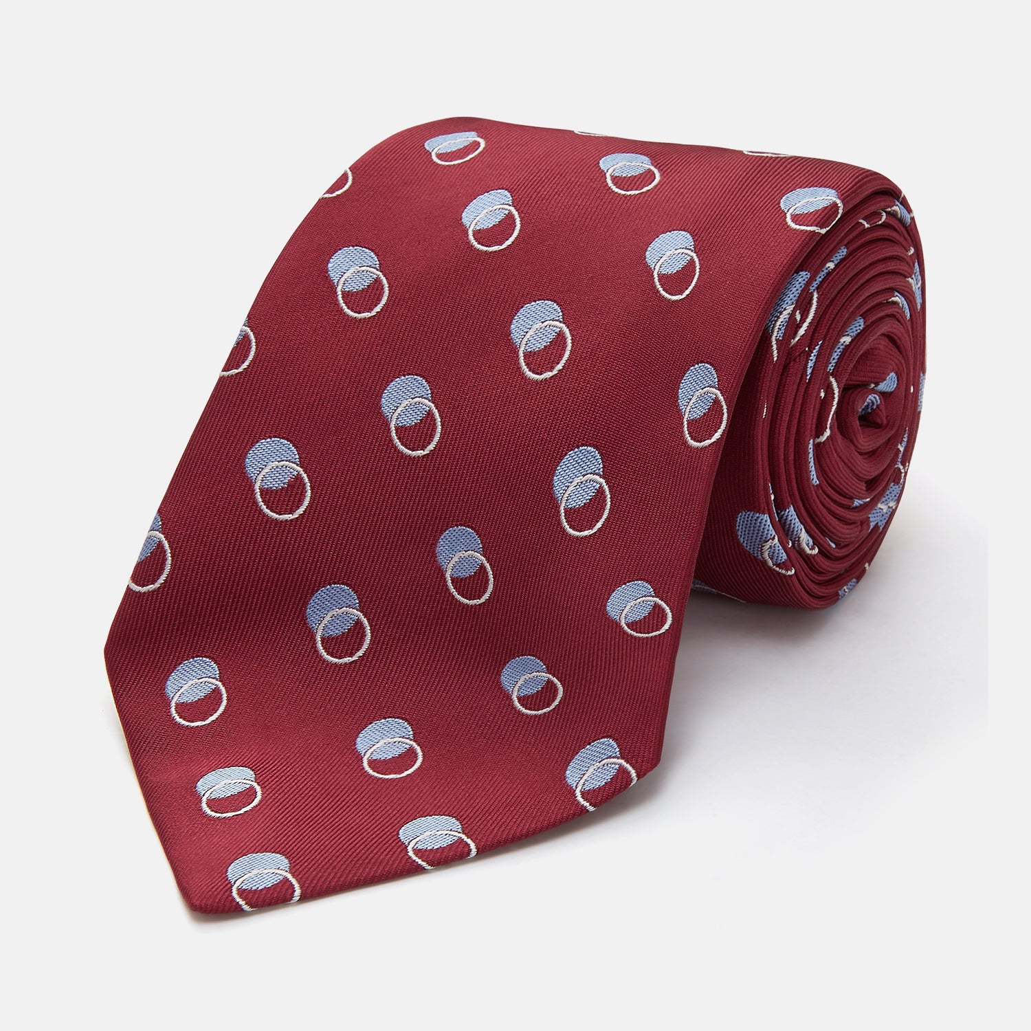 View 1 of product The Turnbull & Asser Red Multi Circle Silk Tie features small blue and white rings on red silk, making it ideal for pairing with bold shirts or adding vibrant flair to summer looks. Shown rolled at one end against a plain white background.