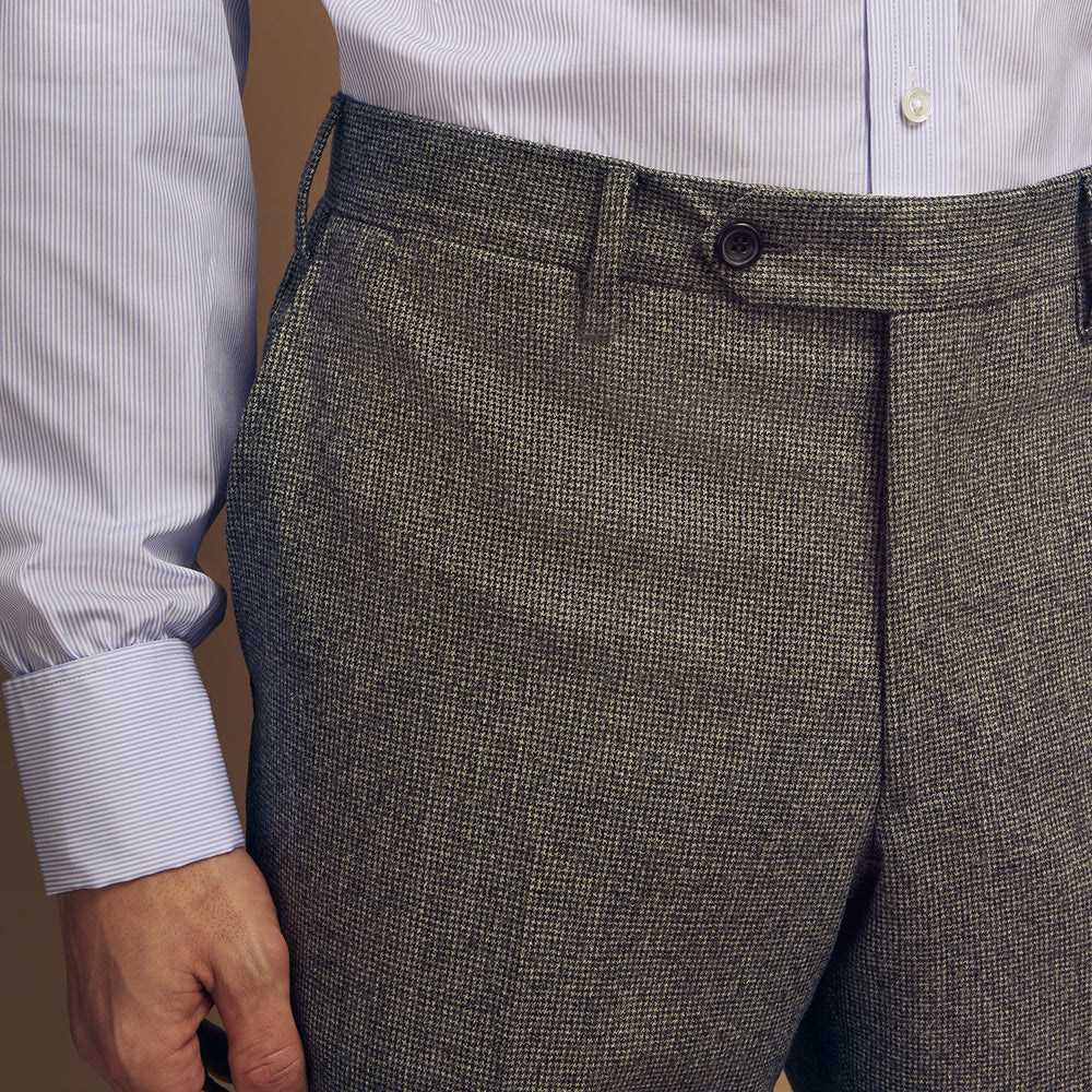 A person wears Turnbull & Asser Grey Puppytooth Wool Manson Trousers, a white dress shirt with rolled-up sleeves, and black shoes, standing against a plain beige background.
