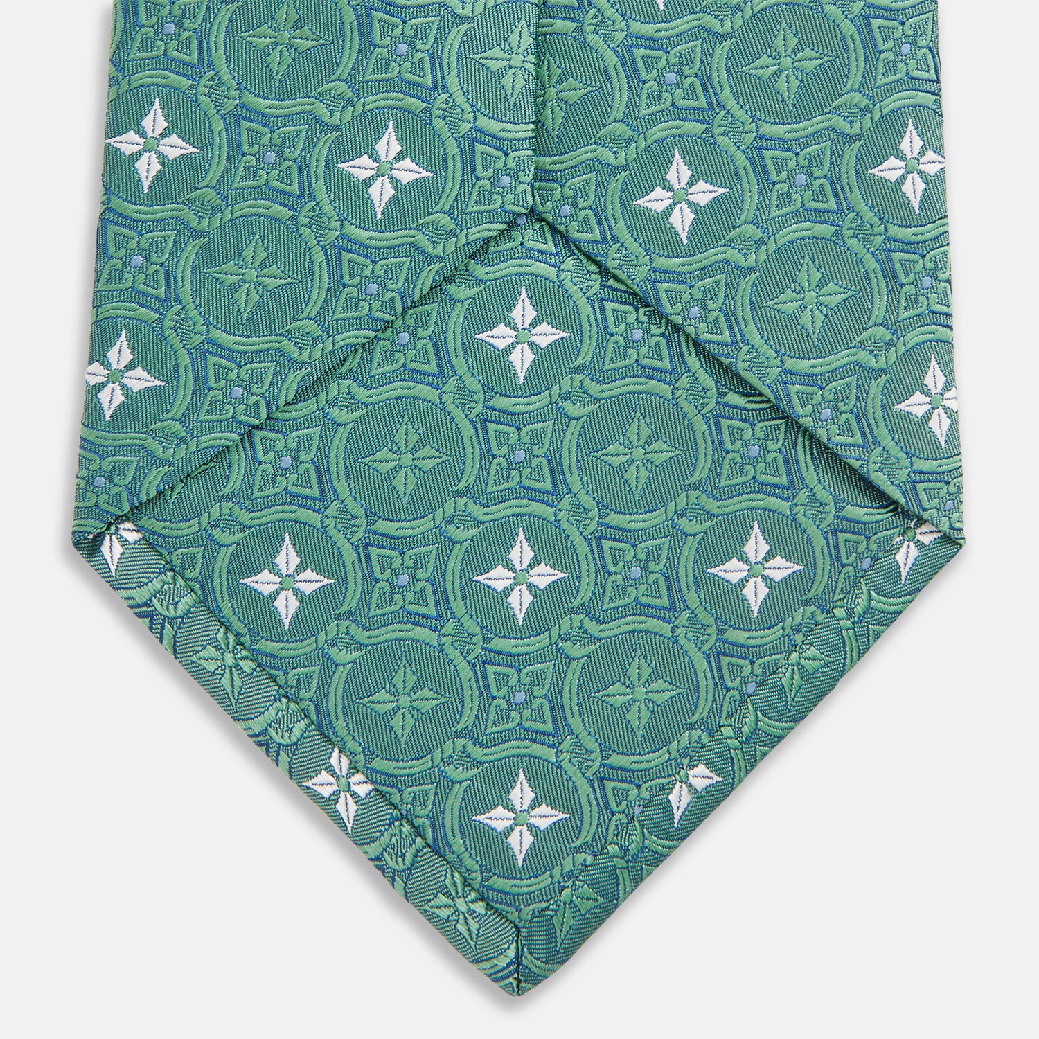 View 4 of product The Turnbull & Asser Green Multi Neat Silk Tie, handmade and featuring a white and light blue geometric pattern, is shown rolled up on a plain white background.