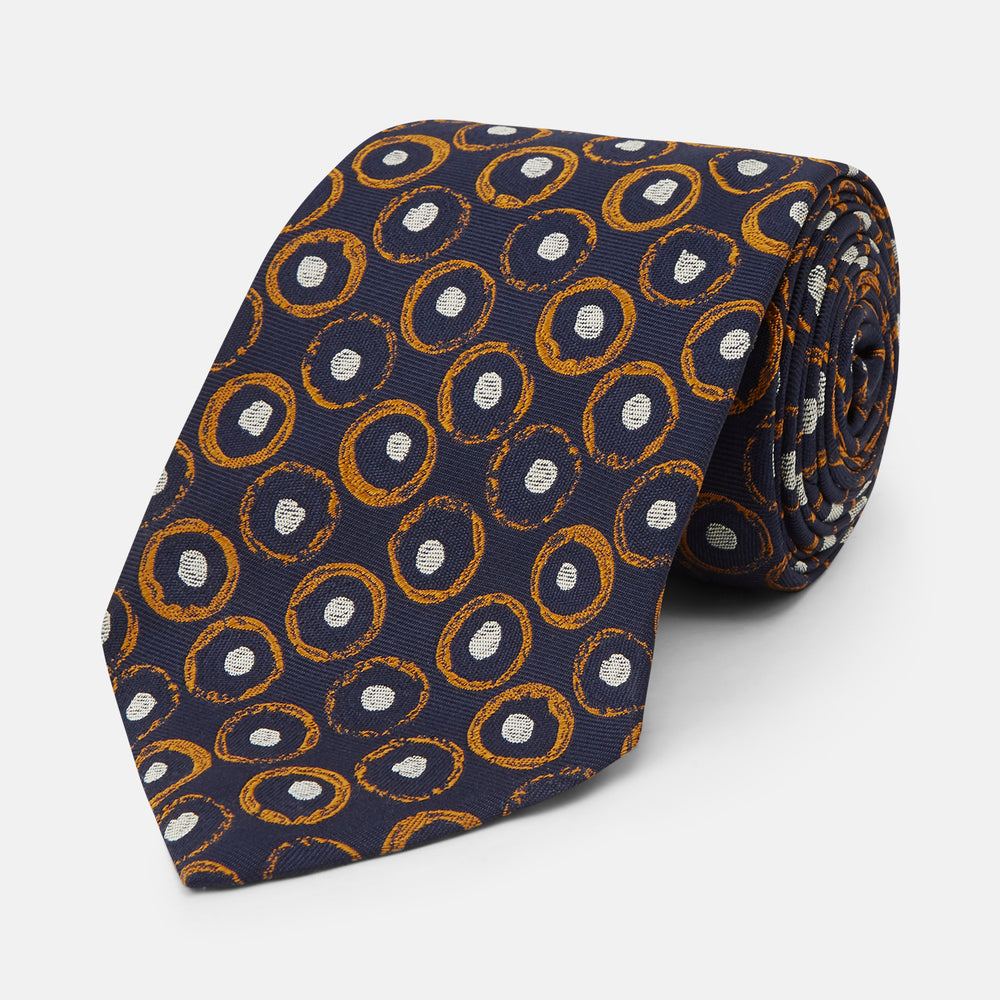 The Turnbull & Asser Orange Multi Circle Printed Silk Tie features navy blue silk with orange circles and white ovals, perfect for elevating any formal outfit. Shown on a plain white background.